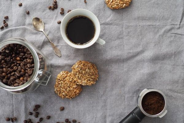 Flat lay of a coffee setup including an elegant cup of black coffee, cookies, and a jar of coffee beans on a linen cloth.