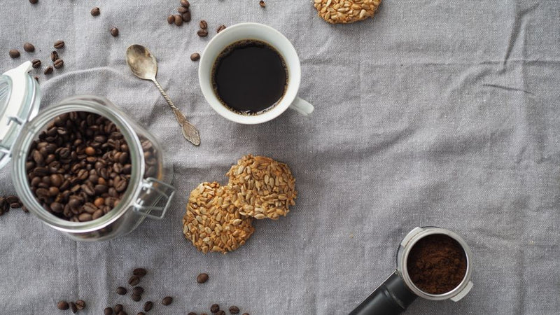 Flat lay of a coffee setup including an elegant cup of black coffee, cookies, and a jar of coffee beans on a linen cloth.
